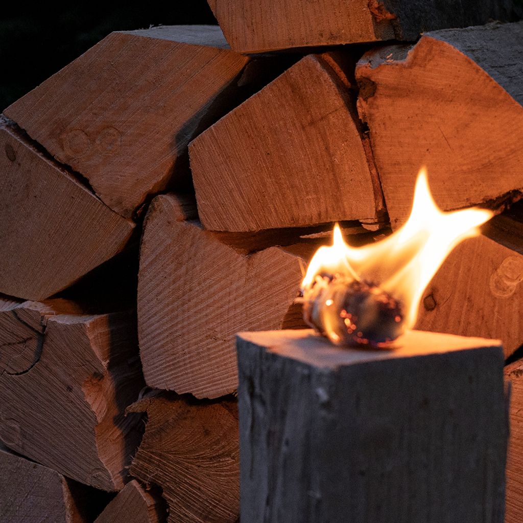 A close-up of a small torch with a flame burning, placed on a wooden block, with a stack of firewood in the background.